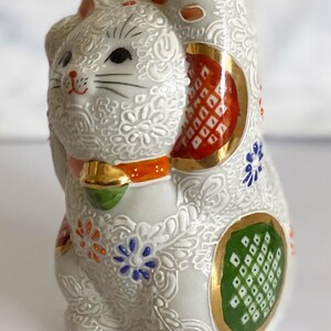 May include: White ceramic Maneki-Neko, or lucky cat, figurine with raised paw. The cat is decorated with intricate floral patterns and colorful accents, including red, orange, green, blue, and gold.