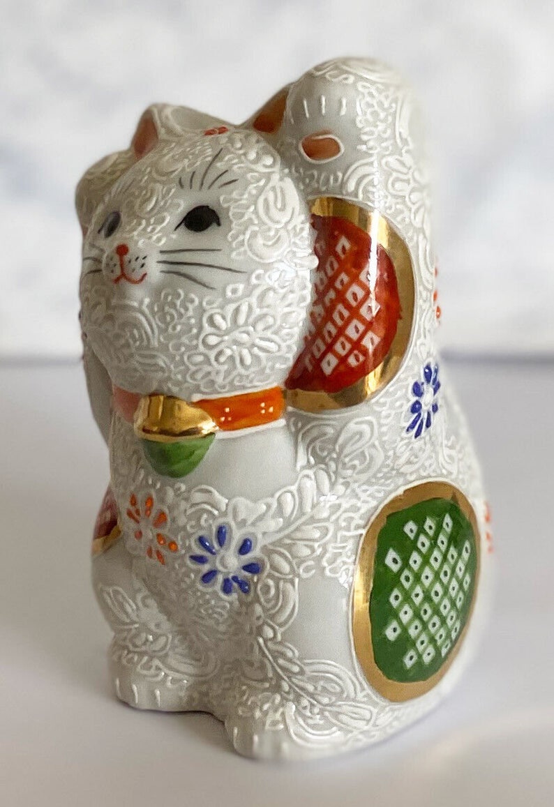 May include: White ceramic Maneki-Neko, or lucky cat, figurine with raised paw. The cat is decorated with intricate floral patterns and colorful accents, including red, orange, green, blue, and gold.
