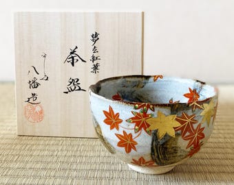 以下が含まれることがあります： 水色とグレーの背景に、赤と金のカエデの葉が描かれた陶器の茶碗。 茶碗の隣には、日本語の文字と赤い印章が書かれた木箱があります。