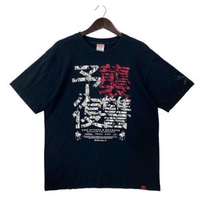 Puede incluir: Camiseta negra de manga corta con un estampado gráfico de caracteres japoneses en blanco y rojo. El texto "PRE-ATTACK & REVENGE MAXIMUM THE HORMONE" está impreso debajo del gráfico.