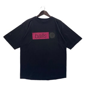 Pode incluir: Camiseta preta com um patch retangular rosa com a palavra "balls" em branco. Um pequeno desenho circular está à direita do patch. A camiseta está pendurada em um cabide de madeira.