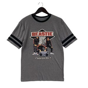 May include: Gray t-shirt with black stripes on the sleeves and a black collar. The shirt features a graphic of the Beastie Boys with the text "BEASTIE BOYS" and "Solid Gold Hits".