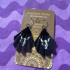 Puede incluir: Pendientes de cara de demonio en blanco y negro hechos con materiales reciclados. Los pendientes tienen forma de concha y tienen un alambre negro envuelto alrededor de la parte superior. Los pendientes están en una tarjeta marrón con el texto "Upcycled Ashley upcycledashley.com Handmade Treasures".