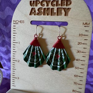 May include: Upcycled earrings made from green and white Heineken beer can tops, with red wire accents. The earrings are displayed on a wooden stand that reads "UPCYCLED ASHLEY" and includes inch and millimeter measurements.
