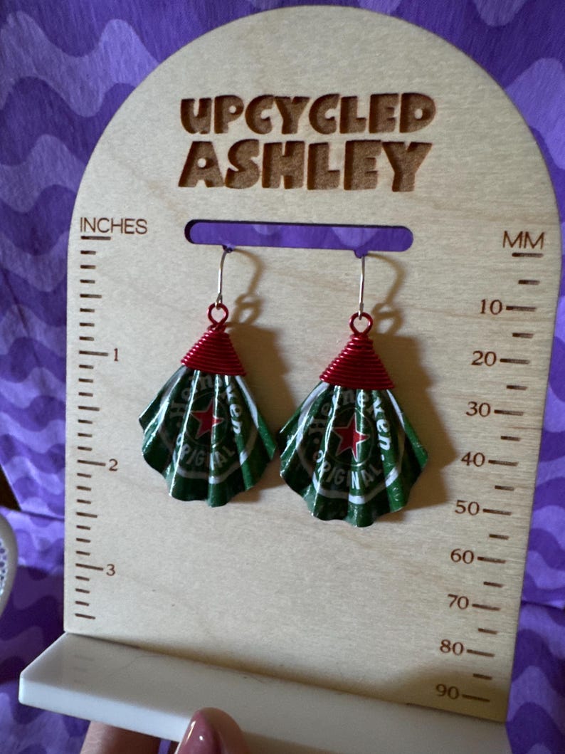 May include: Upcycled earrings made from green and white Heineken beer can tops, with red wire accents. The earrings are displayed on a wooden stand that reads "UPCYCLED ASHLEY" and includes inch and millimeter measurements.