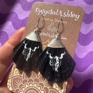 Puede incluir: Pendientes negros con un diseño de demonio cornudo blanco, sujetos a ganchos plateados. Los pendientes tienen forma de cono de alambre plateado. Se exhiben en una tarjeta marrón con el texto "Upcycled Ashley".