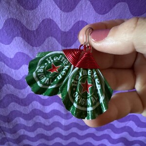 May include: A pair of handmade earrings featuring green and white bottle caps with the text "Heineken Original" and a red star. The caps are attached to silver hooks with red wire wrapping.
