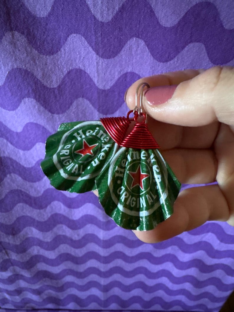 May include: A pair of handmade earrings featuring green and white bottle caps with the text "Heineken Original" and a red star. The caps are attached to silver hooks with red wire wrapping.