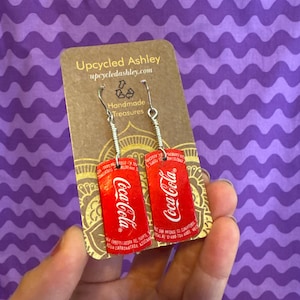 May include: A pair of earrings made from recycled Coca-Cola cans. The earrings are silver and have a red Coca-Cola logo on them. The earrings are on a brown card with the text "Upcycled Ashley" and "upcycledashley.com".