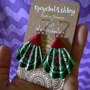 May include: Upcycled earrings made from green and white Heineken beer cans, with red wire accents. The earrings are displayed on a brown card with the text "Upcycled Ashley" and "Trash to Treasure".