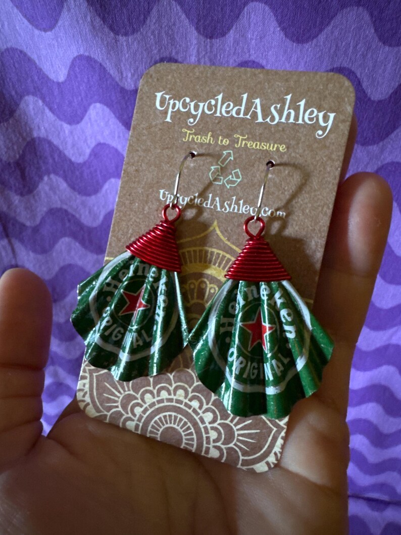 May include: Upcycled earrings made from green and white Heineken beer cans, with red wire accents. The earrings are displayed on a brown card with the text "Upcycled Ashley" and "Trash to Treasure".