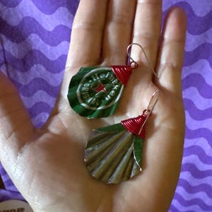 May include: Handmade earrings crafted from recycled Heineken bottle caps. The earrings feature green and silver metal with red wire accents. The design includes the Heineken logo and a star. The earrings are held in a hand.