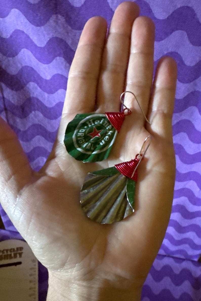 May include: Handmade earrings crafted from recycled Heineken bottle caps. The earrings feature green and silver metal with red wire accents. The design includes the Heineken logo and a star. The earrings are held in a hand.