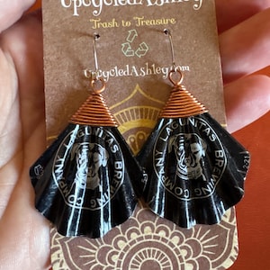 May include: Handmade earrings made from upcycled materials. The earrings feature a black, fan-shaped design with the text "LAGUNITAS BREWING COMPANY" and a dog illustration. The top is wrapped in copper wire. The earrings are attached to a brown card with the text "Upcycled Ashley".