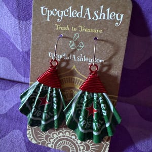May include: Upcycled earrings made from green and white Heineken beer cans, with red wire accents. The earrings are displayed on a brown card with the text "Upcycled Ashley" and "Trash to Treasure".
