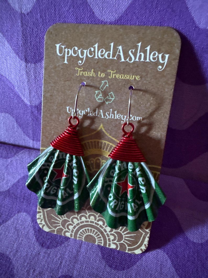 May include: Upcycled earrings made from green and white Heineken beer cans, with red wire accents. The earrings are displayed on a brown card with the text "Upcycled Ashley" and "Trash to Treasure".