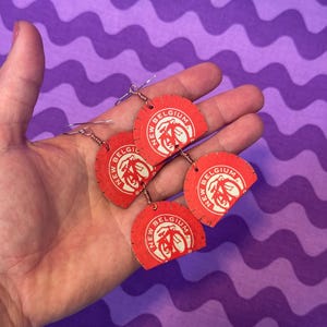 Könnte beinhalten: Drei Paar Ohrringe aus roten und weißen Flaschendeckeln der New Belgium Brewing Company. Jeder Ohrring zeigt ein weißes Fahrradlogo mit den Worten "New Belgium" und "Belgium" in weiß.