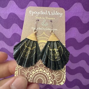Puede incluir: Pendientes negros y dorados hechos a mano con un diseño en forma de abanico. Cada pendiente presenta un alambre dorado y un emblema circular con un diseño detallado. Los pendientes se muestran en una tarjeta marrón con el texto "Upcycled Ashley".