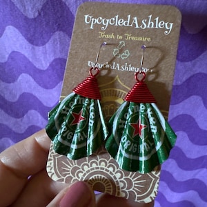 May include: Upcycled earrings made from Heineken beer cans. The earrings have a red wire top and a green and white fan-shaped bottom. The earrings are displayed on a brown card with the text "Upcycled Ashley" and "Trash to Treasure".