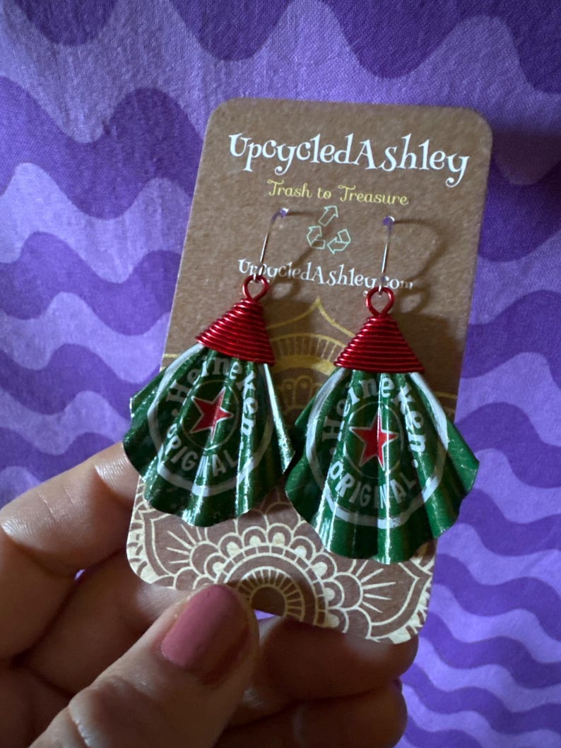 May include: Upcycled earrings made from Heineken beer cans. The earrings have a red wire top and a green and white fan-shaped bottom. The earrings are displayed on a brown card with the text "Upcycled Ashley" and "Trash to Treasure".