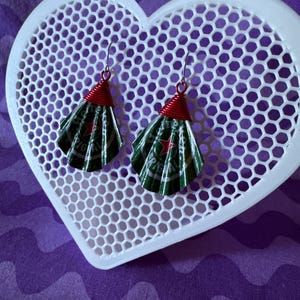 May include: Handmade earrings featuring green and silver fan-shaped elements, crafted from recycled materials. The earrings have red wire accents and silver hooks. They are displayed on a white heart-shaped, honeycomb-patterned tray against a purple background.