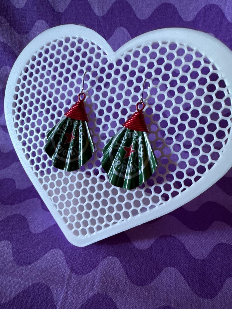 May include: Handmade earrings featuring green and silver fan-shaped elements, crafted from recycled materials. The earrings have red wire accents and silver hooks. They are displayed on a white heart-shaped, honeycomb-patterned tray against a purple background.