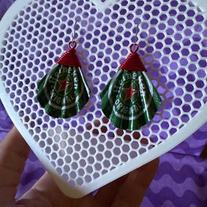 May include: Handmade earrings crafted from green and silver Heineken beer can tops, with red wire accents. The earrings are suspended from silver hooks and are displayed on a white heart-shaped lattice. The background is a purple fabric.
