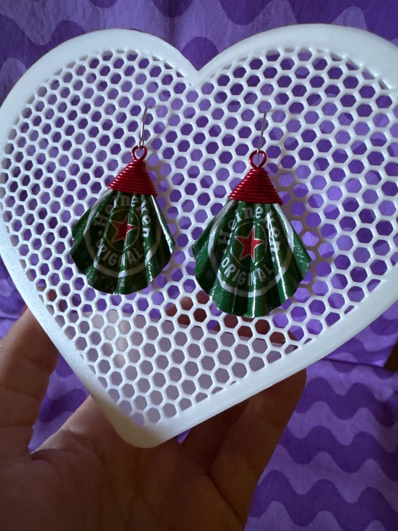 May include: Handmade earrings crafted from green and silver Heineken beer can tops, with red wire accents. The earrings are suspended from silver hooks and are displayed on a white heart-shaped lattice. The background is a purple fabric.