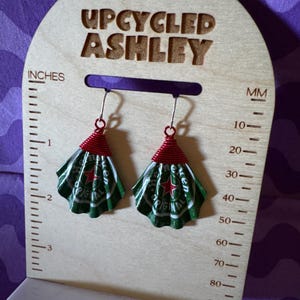 May include: Upcycled earrings made from green and white Heineken beer cans, with red wire accents. The earrings are displayed on a wooden stand that reads "UPCYCLED ASHLEY" and includes inch and millimeter measurements.