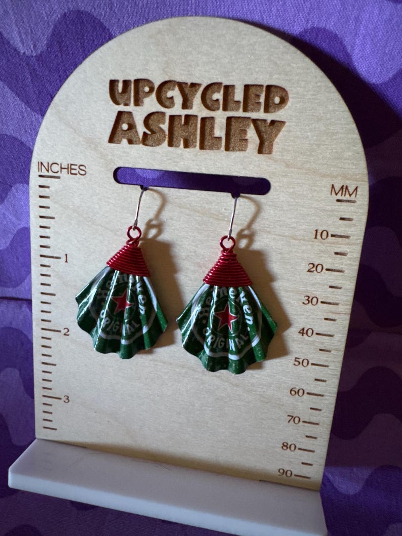 May include: Upcycled earrings made from green and white Heineken beer cans, with red wire accents. The earrings are displayed on a wooden stand that reads "UPCYCLED ASHLEY" and includes inch and millimeter measurements.