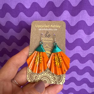 May include: Handmade orange earrings with a sun design, crafted from recycled materials. The earrings have teal wire accents and are displayed on a brown card with the text "Upcycled Ashley" and "Handmade Treasures".