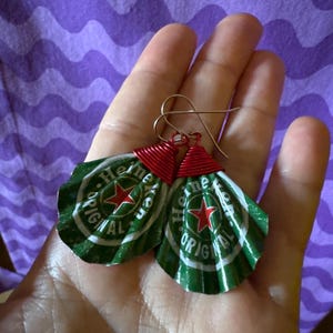 May include: Handmade earrings crafted from Heineken beer bottle caps. The green and white caps feature a red star and the word "Heineken". The caps are crimped and attached to gold-colored ear wires with red wire.