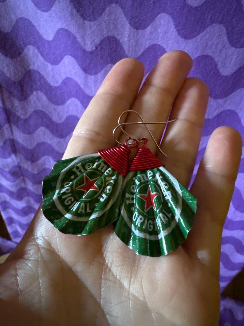 May include: Handmade earrings crafted from Heineken beer bottle caps. The green and white caps feature a red star and the word "Heineken". The caps are crimped and attached to gold-colored ear wires with red wire.
