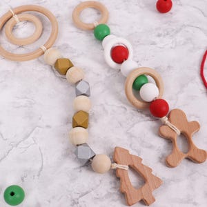 Puede incluir: Juguetes de madera para bebés con anillos, cuentas y formas festivas de madera natural. Un juguete presenta un hombre de jengibre, el otro un árbol de Navidad. Los acentos rojos, verdes y blancos añaden un toque festivo.