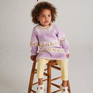 May include: A young child wearing a purple and white striped knitted sweater dress with a floral pattern. The dress is chunky knit and has a round neckline. The child is sitting on a wooden stool and wearing white shoes.