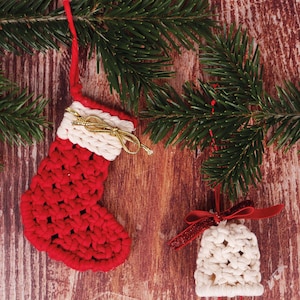 May include: Two handmade Christmas decorations: a red crocheted stocking with a white cuff and gold bow, and a white crocheted bell with a red ribbon and small gold bell. They are displayed with evergreen branches.