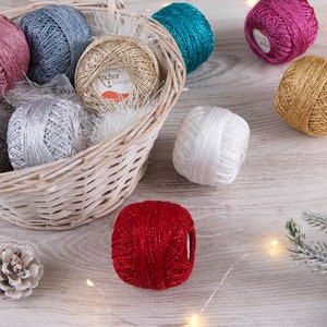 Puede incluir: Una cesta de mimbre llena de ovillos de hilo brillante en varios colores, incluyendo plateado, dorado, rojo, rosa, turquesa y blanco. La cesta está sentada sobre una superficie de madera con ramas de pino y piñas.