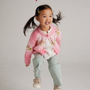 May include: A young girl wearing a pink and white cardigan with a floral pattern jumps in the air. The cardigan is a knit pattern with a button closure. The girl is wearing light blue trousers and beige trainers. The text "Baby Blossom Chunky 1-4 years" is at the bottom of the image.