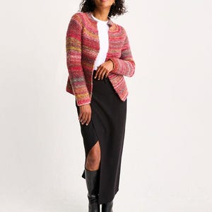 Puede incluir: Una mujer con una chaqueta de punto multicolor, una camiseta blanca y una falda negra con una abertura en el muslo. También lleva botas negras hasta la rodilla. La chaqueta tiene tonos de rosa, rojo, amarillo y verde.