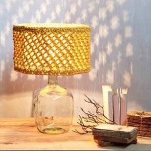 May include: A table lamp with a crocheted lampshade made of natural fiber rope. The lampshade is a light yellow color and has a woven pattern. The lamp base is a clear glass bottle.