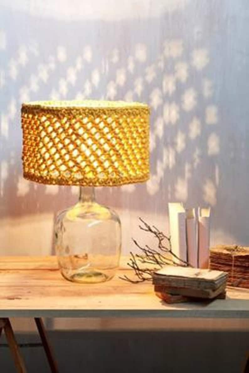 May include: A table lamp with a crocheted lampshade made of natural fiber rope. The lampshade is a light yellow color and has a woven pattern. The lamp base is a clear glass bottle.