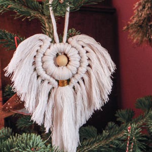 May include: A white macrame angel Christmas decoration. The angel has a circular head with a wooden bead centre, a gold-coloured metal ring, and fringed wings. It hangs from a white cord and is displayed on a Christmas tree.