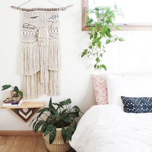 May include: A white macrame wall hanging with a wood dowel at the top, a potted plant, and a white bed with a pink and white patterned pillow, a white pillow, and a blue and white patterned pillow.