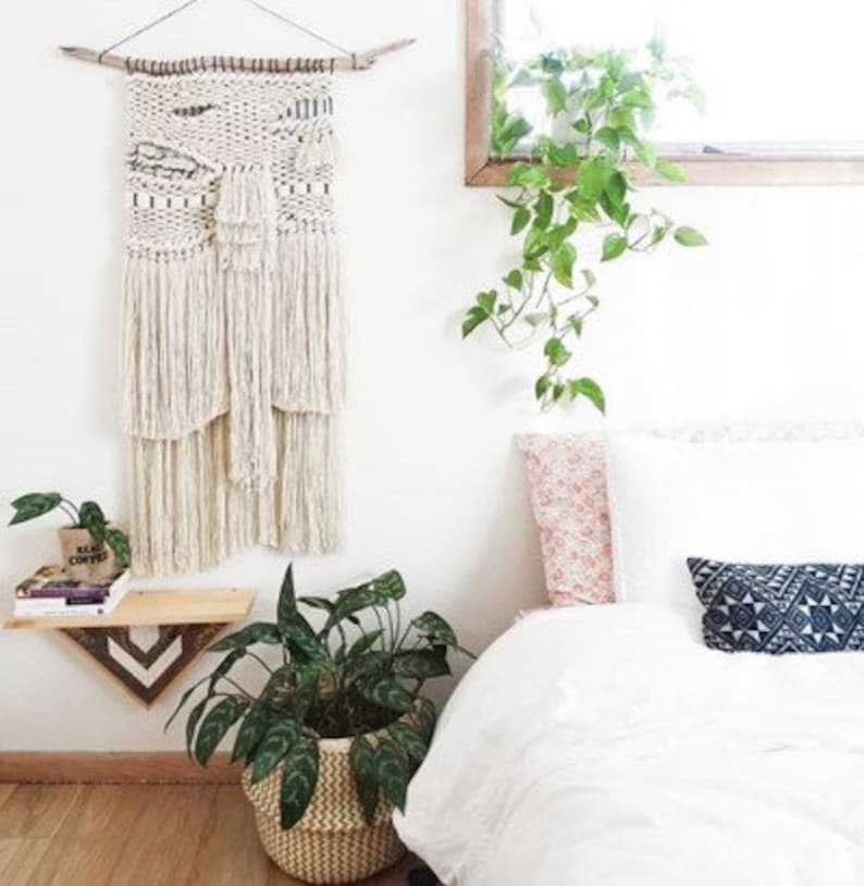 May include: A white macrame wall hanging with a wood dowel at the top, a potted plant, and a white bed with a pink and white patterned pillow, a white pillow, and a blue and white patterned pillow.