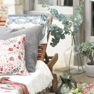 May include: A white lounge chair with a gray pillow and a floral patterned pillow. The chair is on a patio with a vase of eucalyptus branches and a green pitcher with white flowers.