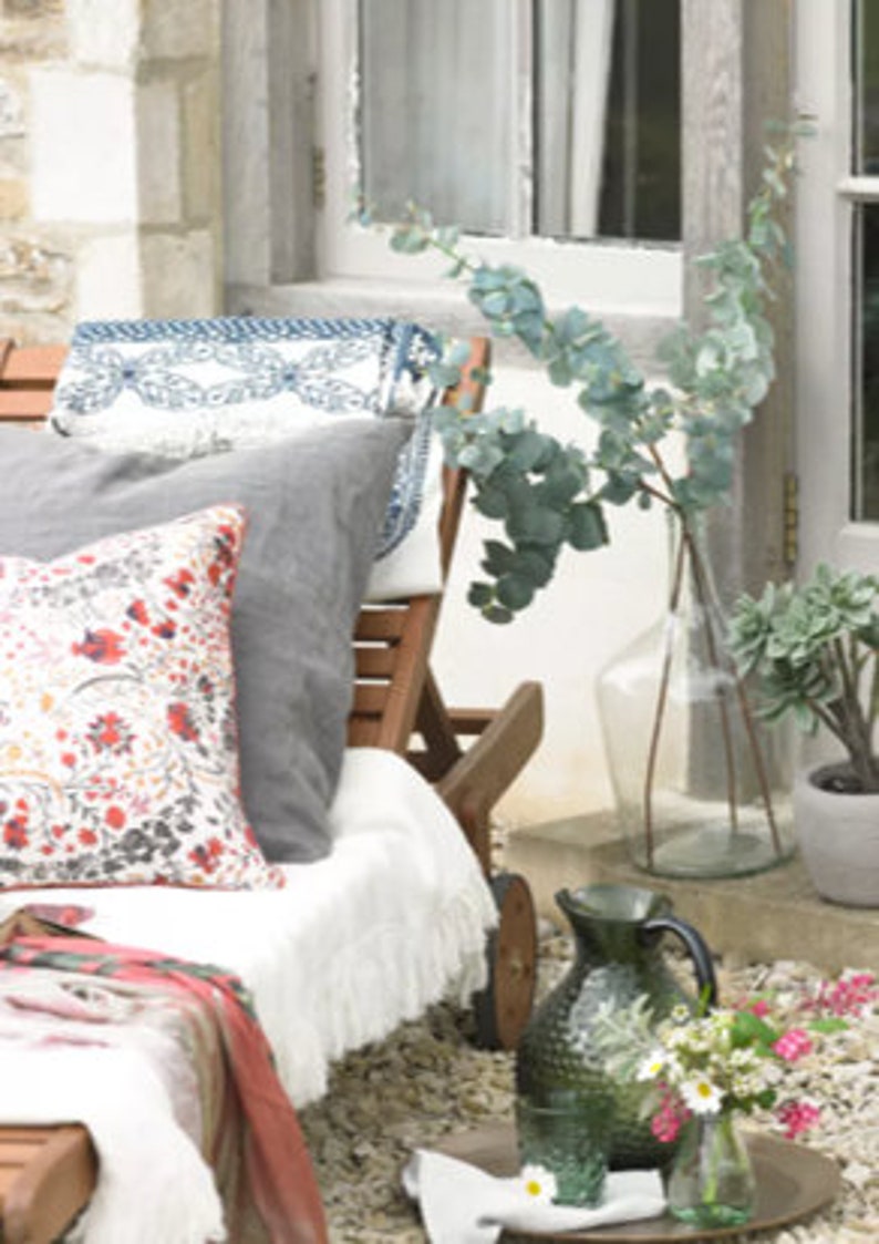 May include: A white lounge chair with a gray pillow and a floral patterned pillow. The chair is on a patio with a vase of eucalyptus branches and a green pitcher with white flowers.