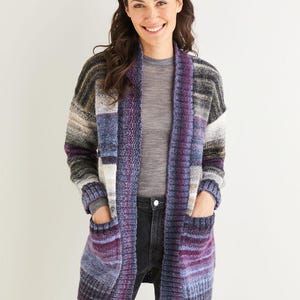 May include: A long, open-front cardigan in a color block design. The sweater features shades of purple, blue, gray, and cream. It has two front pockets and a shawl collar. The cardigan is made of a thick, textured knit.