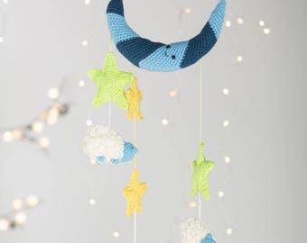 May include: A blue and white crocheted crescent moon mobile with three white sheep and four yellow and green stars hanging from it.