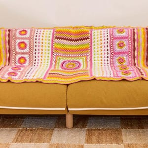 Peut inclure: Une couverture en crochet rose, jaune, verte et marron avec différents motifs, notamment des cœurs, des fleurs et des carrés. La couverture est posée sur un canapé jaune moutarde.