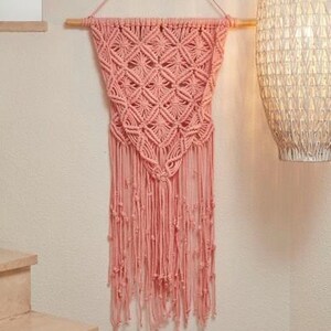 May include: A pink macrame wall hanging with a geometric pattern and fringe. The hanging is made of cotton cord and is attached to a wooden dowel.